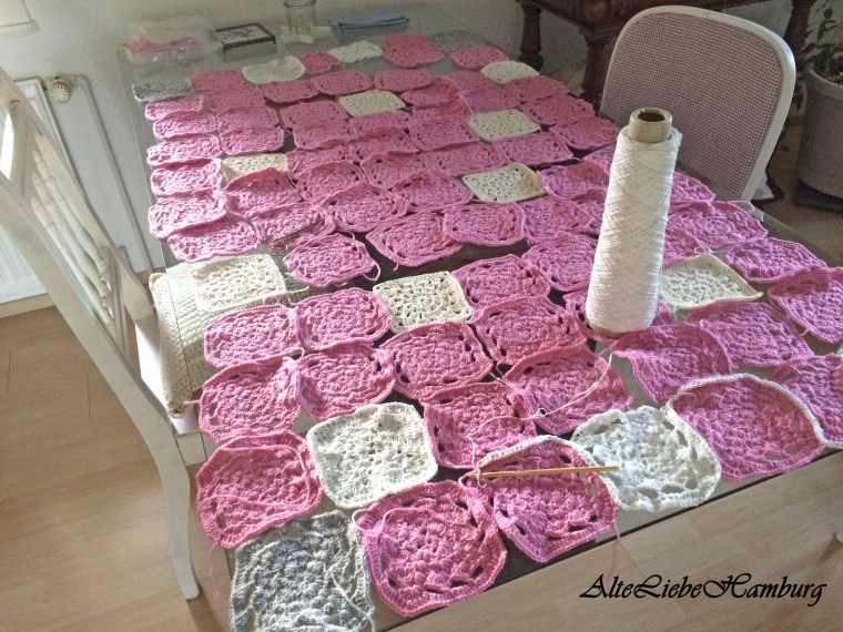 Viele Vierecke zum Zusammennähen auf einem Tisch liegend. Eine Granny Square Decke in rose, weiss, grau