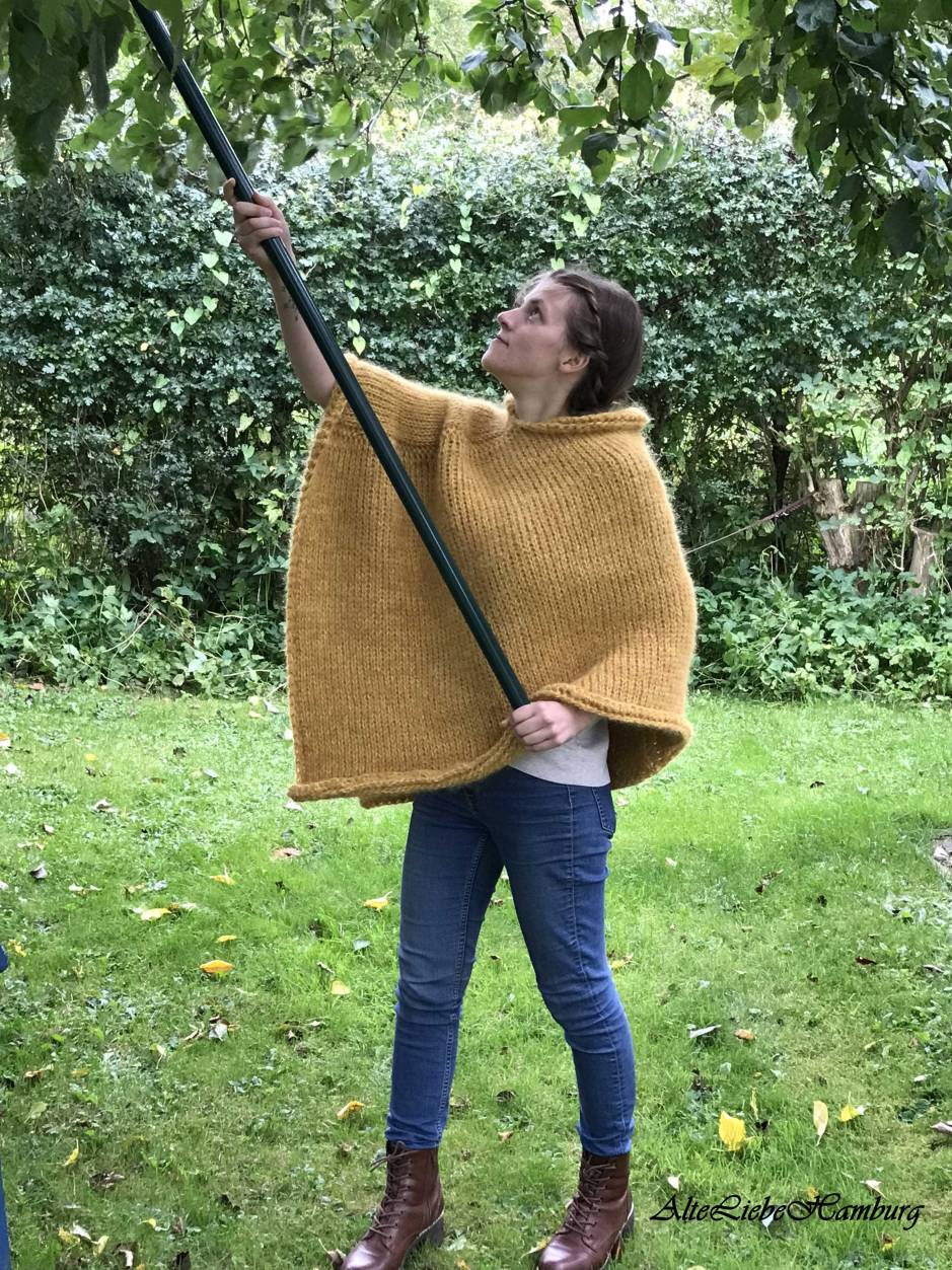 Junge Frau pflückt Äpfel. Trägt einen zitronensenfgelben Poncho aus Alpaca Wolle